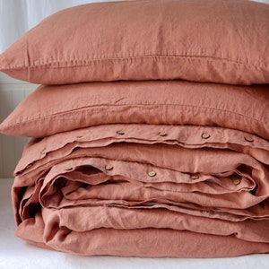 May include: A stack of terracotta-colored linen bedding, including two pillowcases and a folded duvet cover. The duvet cover is secured with wooden buttons. The bedding is neatly arranged on a white surface, suggesting a bedroom setting.