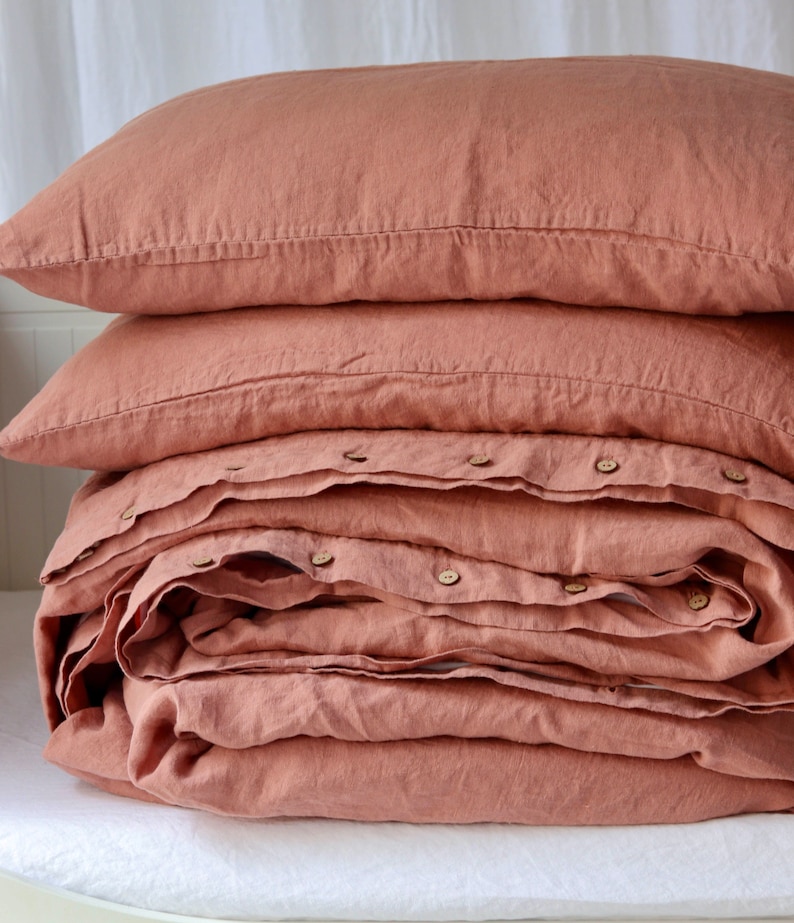 May include: A stack of terracotta-colored linen bedding, including two pillowcases and a folded duvet cover. The duvet cover is secured with wooden buttons. The bedding is neatly arranged on a white surface, suggesting a bedroom setting.