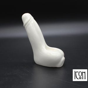 May include: A white ceramic sculpture of a stylized phallus. The sculpture is smooth and glossy, with a rounded base and a detailed head. The background is black, and the sculpture is lit from above. The logo "icon" is in the bottom right corner.