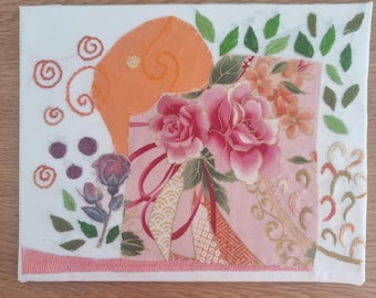May include: A fabric art piece featuring a floral design with pink and peach roses, green leaves, and orange accents. The artwork is on a white canvas with embroidered details and a decorative border.