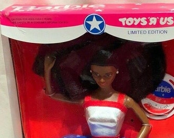 Könnte beinhalten: Eine schwarze Barbie-Puppe, die ein rot-weiß-blaues Kleid mit einem silbernen Sternmuster trägt. Die Puppe hält einen Wahlkampfbutton mit der Aufschrift "Barbie for President". Die Schachtel ist mit "Toys 'R' Us Limited Edition" beschriftet.