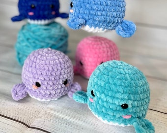 Könnte beinhalten: Fünf gehäkelte Wal-Plüschtiere in verschiedenen Farben: Blau, Lila, Rosa, Türkis und Hellblau. Die Wale haben weiße Bäuche und schwarze Augen.