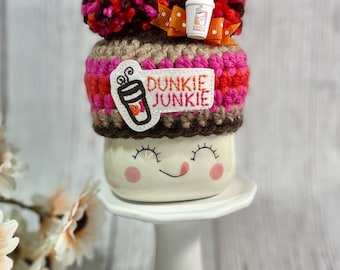 May include: A whimsical coffee mug with a crocheted cozy featuring the text "Dunkie Junkie." The cozy is adorned with pink, orange, and brown yarn, topped with pom-poms and a small bow. The mug has a smiling face design.