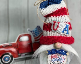 Könnte beinhalten: Patriotischer Zwerg mit einer rot-weiß-blauen Strickmütze mit dem Wort "USA" darauf. Der Zwerg hält ein rundes Holzschild mit der Aufschrift "Glaube Familie Freiheit".
