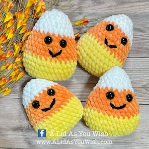 May include: Four plush, smiling candy corn decorations. Each has a white top, orange middle, and yellow bottom. Black button eyes and a stitched black smile complete the design. The image includes the text "A Lid As You Wish" and a website address.