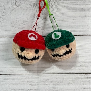May include: Two crocheted plush characters, one red and one green, with a white face and a black smile. They are hanging from a string with a metal clip.