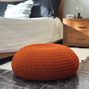 Op de afbeelding: Een ronde, oranje gehaakte poef, een decoratief en functioneel meubelstuk. De poef is gemaakt van dik, getextureerd garen en staat op een donkere tegelvloer. De achtergrond omvat een bed met kussens en een houten nachtkastje.