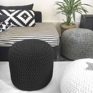May include: A modern living room scene featuring a black, hand-crocheted ottoman. Other ottomans in gray and white are also visible. A black and white striped daybed and patterned pillows add to the contemporary decor. A potted plant and woven basket complete the scene.