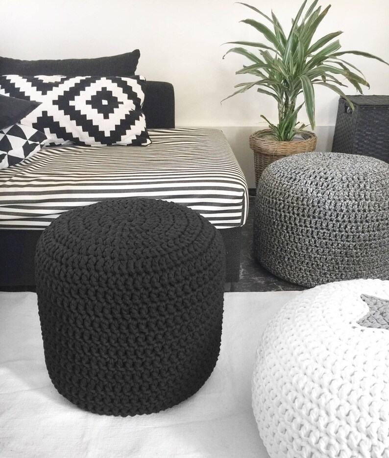 May include: A modern living room scene featuring a black, hand-crocheted ottoman. Other ottomans in gray and white are also visible. A black and white striped daybed and patterned pillows add to the contemporary decor. A potted plant and woven basket complete the scene.