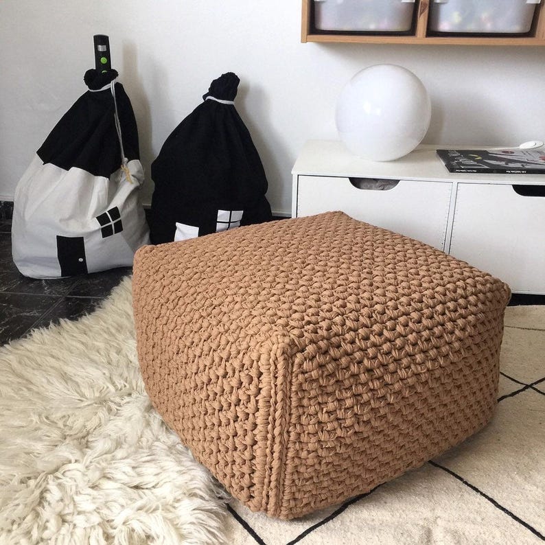 May include: A square, tan-colored, crocheted ottoman sits on a white and black patterned rug. Two house-shaped storage bags, one black and one white, are in the background. A white cabinet and a white sphere are also visible.