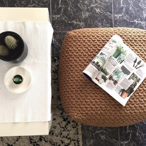 May include: Overhead shot of a living room scene with a white table, a woven pouf, and a black tiled floor. A potted cactus and a small ceramic dish with a plant sit on the table. A magazine about plants rests on the pouf.