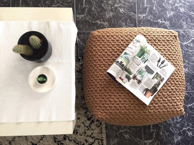 May include: Overhead shot of a living room scene with a white table, a woven pouf, and a black tiled floor. A potted cactus and a small ceramic dish with a plant sit on the table. A magazine about plants rests on the pouf.