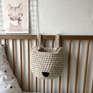 Può includere: Un cestino portaoggetti all'uncinetto beige a forma di muso d'orso, con naso nero e dettagli con perline di legno. Il cestino è appeso a una culla di legno, con un'illustrazione di volpe sullo sfondo. Per la cameretta.
