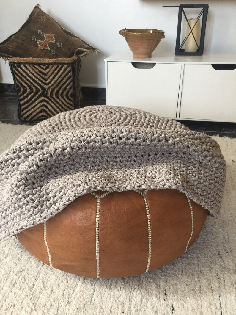 May include: A brown leather pouf with white stitching has a taupe crocheted cover draped over the pouf.