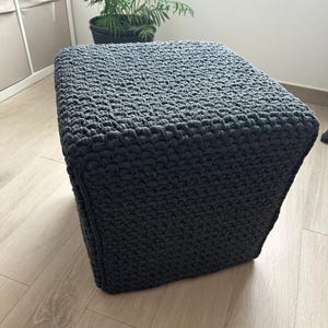 May include: A square, dark gray crocheted ottoman. The textured surface is made of thick yarn, creating a dense, solid appearance. The ottoman is placed on a light-colored wooden floor, with a potted plant visible in the background.