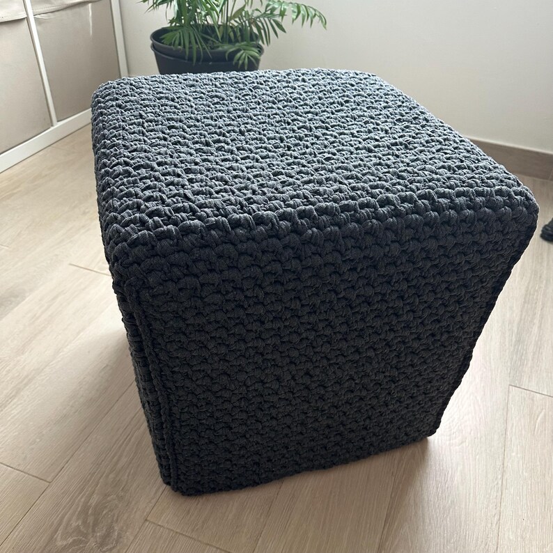 May include: A square, dark gray crocheted ottoman. The textured surface is made of thick yarn, creating a dense, solid appearance. The ottoman is placed on a light-colored wooden floor, with a potted plant visible in the background.