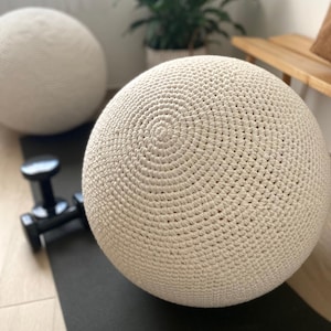 May include: Two cream-colored, crocheted exercise balls are featured in this image. The balls have a spiral pattern at the top and are sitting on a black exercise mat. A black dumbbell is in the foreground. A plant in a woven basket is in the background.
