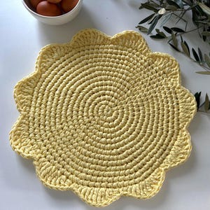 May include: A round, pale yellow crochet mat featuring a textured, spiraling stitch pattern and a scalloped, petal-like edge. The circular textile piece is crafted from thick yarn and displays a consistent, dense weave throughout its entire surface.