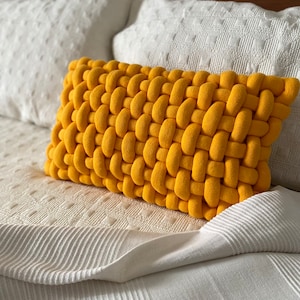 May include: A rectangular, mustard-yellow decorative pillow with a woven design. The pillow is placed on a white bed with white pillows in the background. The woven texture is prominent, creating a cozy and textured appearance.