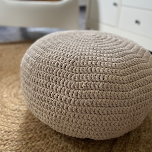 May include: A large, round, beige crocheted pouffe or ottoman. The pouffe is made with a thick, textured yarn and has a simple, classic design.