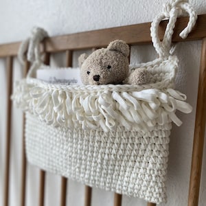 May include: A cream-colored crocheted hanging storage basket with a fringed top edge, featuring a textured stitch pattern. A small, light brown plush teddy bear sits inside the basket, which is suspended by two knotted fabric ties.