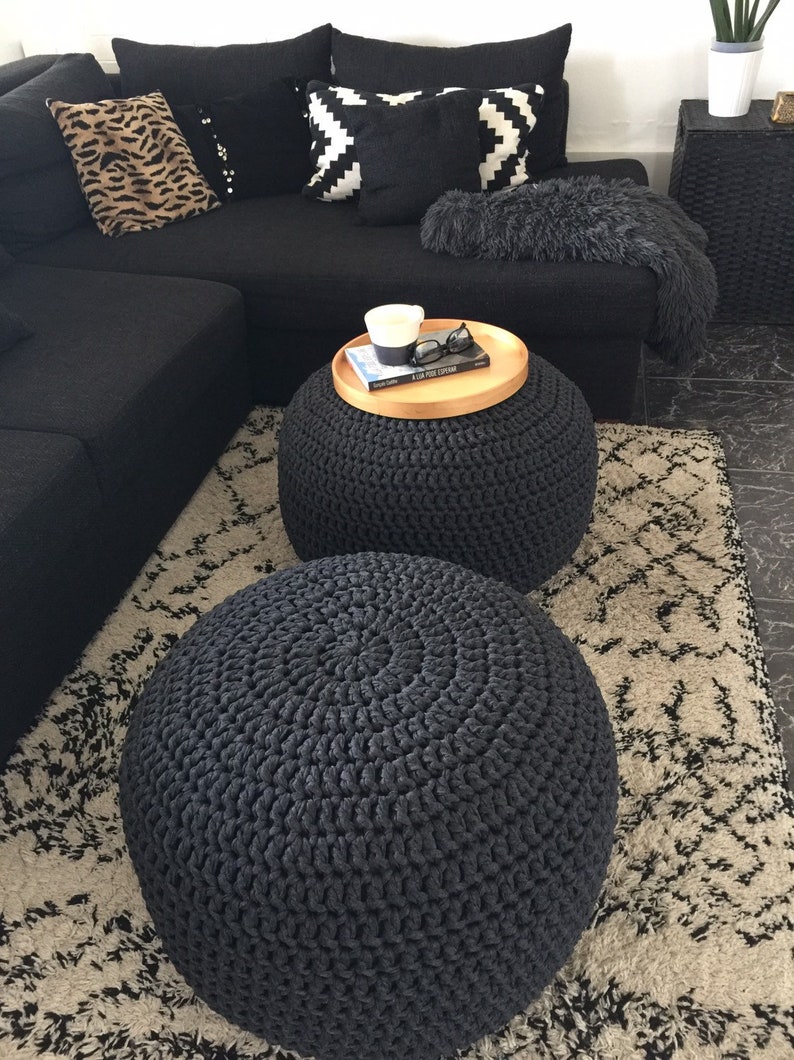 May include: A modern living room setup featuring a black sectional sofa with decorative pillows in various patterns. Two dark gray, round, knitted ottomans sit on a patterned rug. A wooden tray holds a candle, book, and glasses.