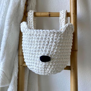 May include: A white crocheted hanging basket featuring two small rounded ears at the top rim and a single black oval nose detail in the center. The basket is constructed from thick yarn with a visible stitch pattern and includes two integrated handles.