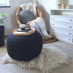 A dark gray round knitted ottoman with a chunky textured surface. A wood trail is on it with a mug and a book. In a corner has a bamboo armchair with a striped blanket and a gray round pillow.