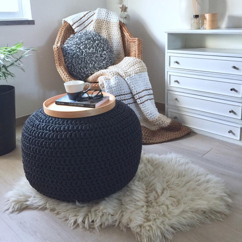 A dark gray round knitted ottoman with a chunky textured surface. A wood trail is on it with a mug and a book. In a corner has a bamboo armchair with a striped blanket and a gray round pillow.