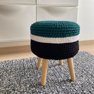 Könnte beinhalten: Ein handgefertigter, runder Hocker mit einem dreibeinigen Holzsockel. Die Oberseite ist dunkelgrün, mit einem weißen Band und einem schwarzen Sockel. Der Hocker besteht aus einem dicken, gestrickten Material. Der Hocker steht auf einem grau-weiß gemusterten Teppich.
