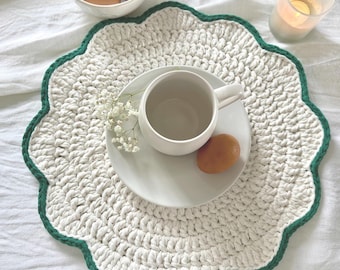 May include: A round, cream-colored crocheted placemat with a scalloped dark green border. Centered on the placemat is a white ceramic mug resting on a matching white saucer, accompanied by a single brown egg and a small sprig of white baby's breath.