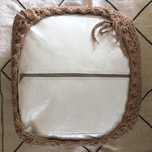 May include: Square, off-white cushion with a tan, crocheted border. The cushion has a zipper across the middle and a braided tie on the side. The crocheted border is thick and adds texture to the design.