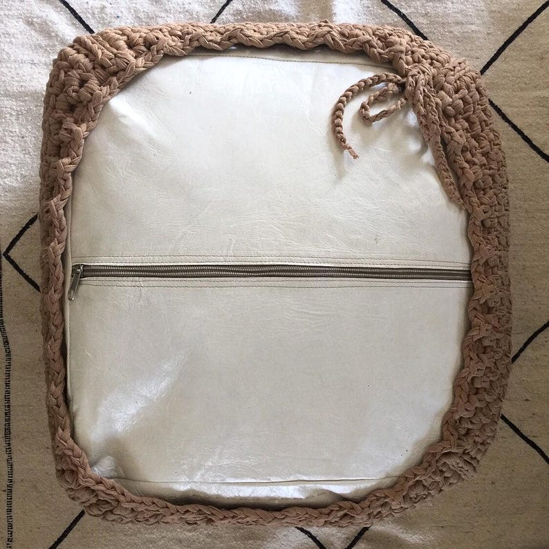 May include: Square, off-white cushion with a tan, crocheted border. The cushion has a zipper across the middle and a braided tie on the side. The crocheted border is thick and adds texture to the design.