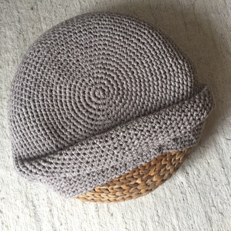 Może przedstawiać: Czapka beanie w kolorze szarobrązowym, szydełkowana, z wywiniętym brzegiem. Czapka wykonana jest z miękkiej, teksturowanej przędzy i ma okrągły kształt. Czapka spoczywa na wiklinowym koszu.