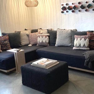 May include: A dark blue sectional sofa with various decorative pillows in shades of gray, cream, and burgundy. A matching ottoman with a tray and magazines sits in front of the sofa. A small side table and a hanging light fixture are also visible.