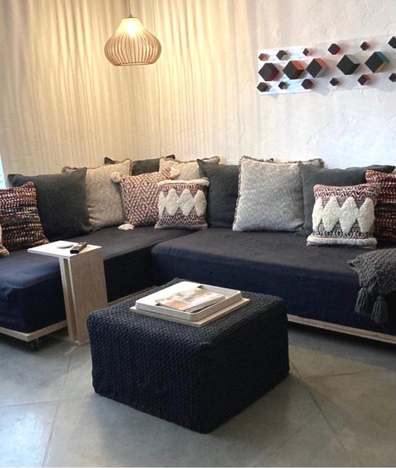 May include: A dark blue sectional sofa with various decorative pillows in shades of gray, cream, and burgundy. A matching ottoman with a tray and magazines sits in front of the sofa. A small side table and a hanging light fixture are also visible.