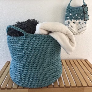 May include: A large, teal-colored, crocheted basket filled with a fluffy, white blanket and a dark gray pillow. A smaller, matching crocheted bag hangs on the wall, with glasses inside. The basket sits on a wooden slatted surface.