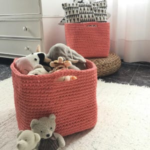 May include: Coral-colored, crocheted storage baskets of various sizes, filled with stuffed animals, sit on a white rug. A black and white patterned pillow rests on top of a basket. The baskets have handles for easy carrying.