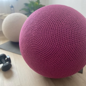 Puede incluir: Dos grandes bolas de ejercicio esféricas de ganchillo. Una es de color magenta intenso y la otra de color crema claro. Las bolas están sobre una esterilla de ejercicio gris, con una pequeña mancuerna en primer plano. Las bolas parecen ser para yoga o pilates.