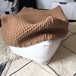 May include: A large, tan-colored, knitted pouf or floor cushion. The knitted top is draped over a white, square-shaped base. The pouf is placed on a patterned rug with a white and black geometric design.