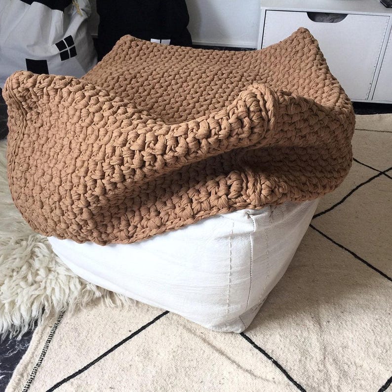 May include: A large, tan-colored, knitted pouf or floor cushion. The knitted top is draped over a white, square-shaped base. The pouf is placed on a patterned rug with a white and black geometric design.