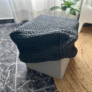 May include: A dark gray, square ottoman with a textured, crocheted cover. The ottoman sits on a light gray base, and is placed on a patterned tile floor. A round, woven rug is visible in the background.