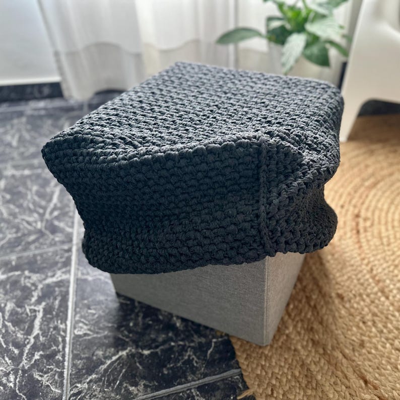 May include: A dark gray, square ottoman with a textured, crocheted cover. The ottoman sits on a light gray base, and is placed on a patterned tile floor. A round, woven rug is visible in the background.