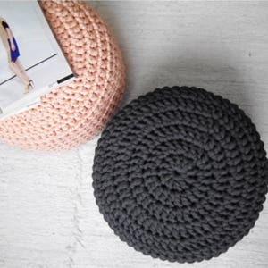 May include: Two round, handmade crochet poufs. One is a soft pink color, and the other is dark gray. A magazine rests on the pink pouf. The poufs are on a white, textured surface.
