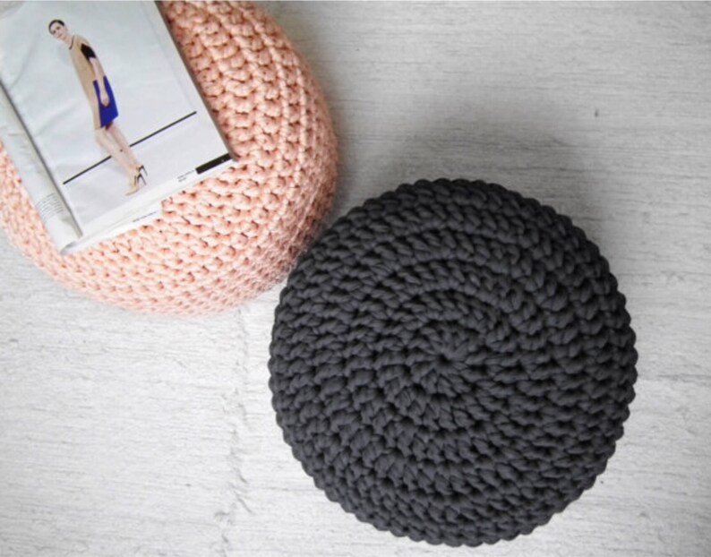 May include: Two round, handmade crochet poufs. One is a soft pink color, and the other is dark gray. A magazine rests on the pink pouf. The poufs are on a white, textured surface.