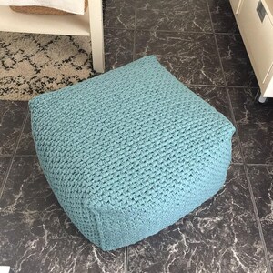 May include: A square, light blue pouf with a textured, woven appearance. The pouf is a solid color and sits on a dark, patterned tile floor. The pouf is a decorative and functional piece of furniture.
