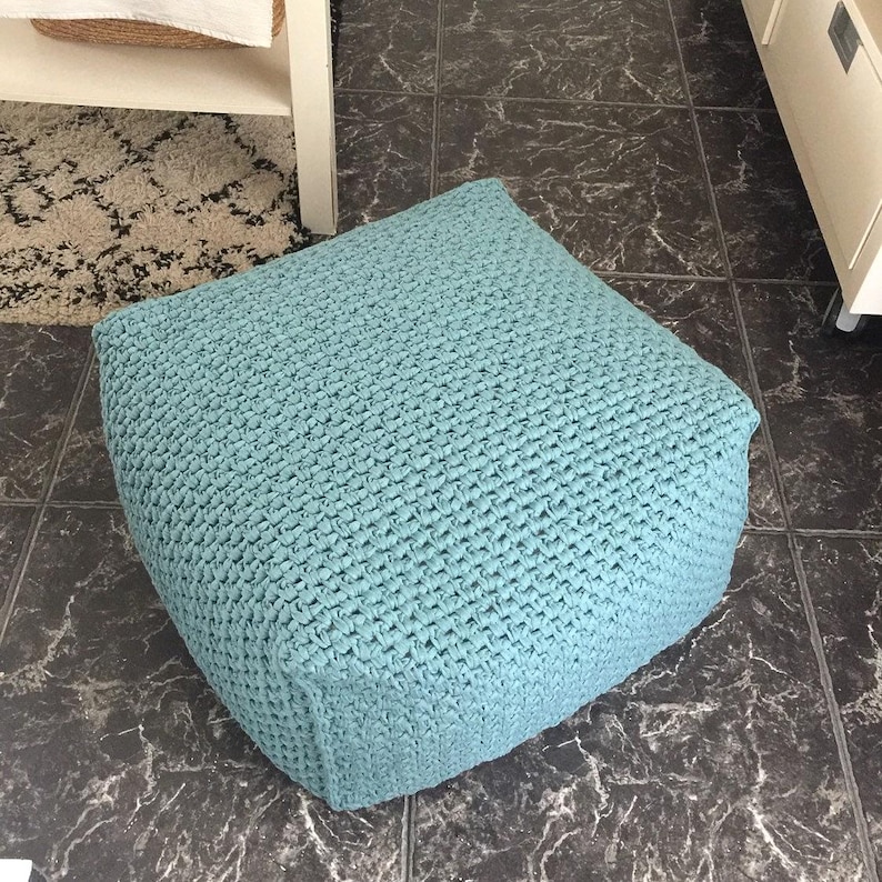May include: A square, light blue pouf with a textured, woven appearance. The pouf is a solid color and sits on a dark, patterned tile floor. The pouf is a decorative and functional piece of furniture.