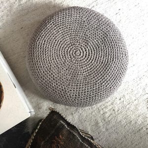 Może przedstawiać: Okrągły, szary puf zrobiony na szydełku ze spiralnym wzorem. Puf wykonany jest z teksturowanej przędzy i leży na jasnej powierzchni. Częściowo widoczna jest poduszka dekoracyjna i książka.