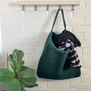 May include: A dark green, handmade crochet tote bag hanging on a white brick wall. The bag is filled with a black hoodie and a black and white striped scarf. The bag has a long handle.