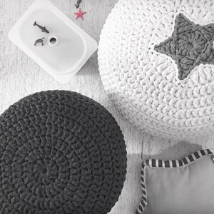 May include: Overhead shot of a white, textured, crocheted pouf with a gray star, a dark gray crocheted pouf, a small gray pillow with striped trim, and a white container with toy fish. The items are on a light-colored surface.